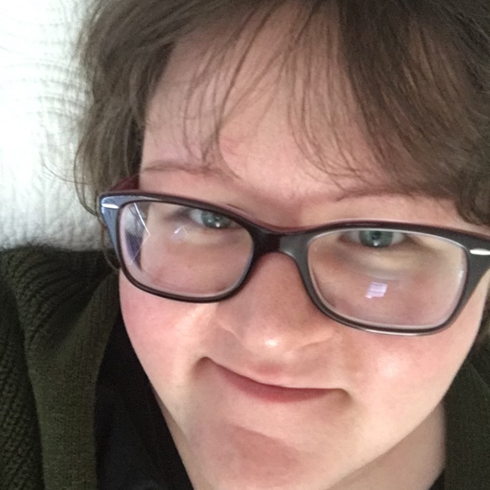 For Screen Readers: A photo of Mia Brody, smiling up at the camera from bed. She's built most of her writing career while lying in bed.